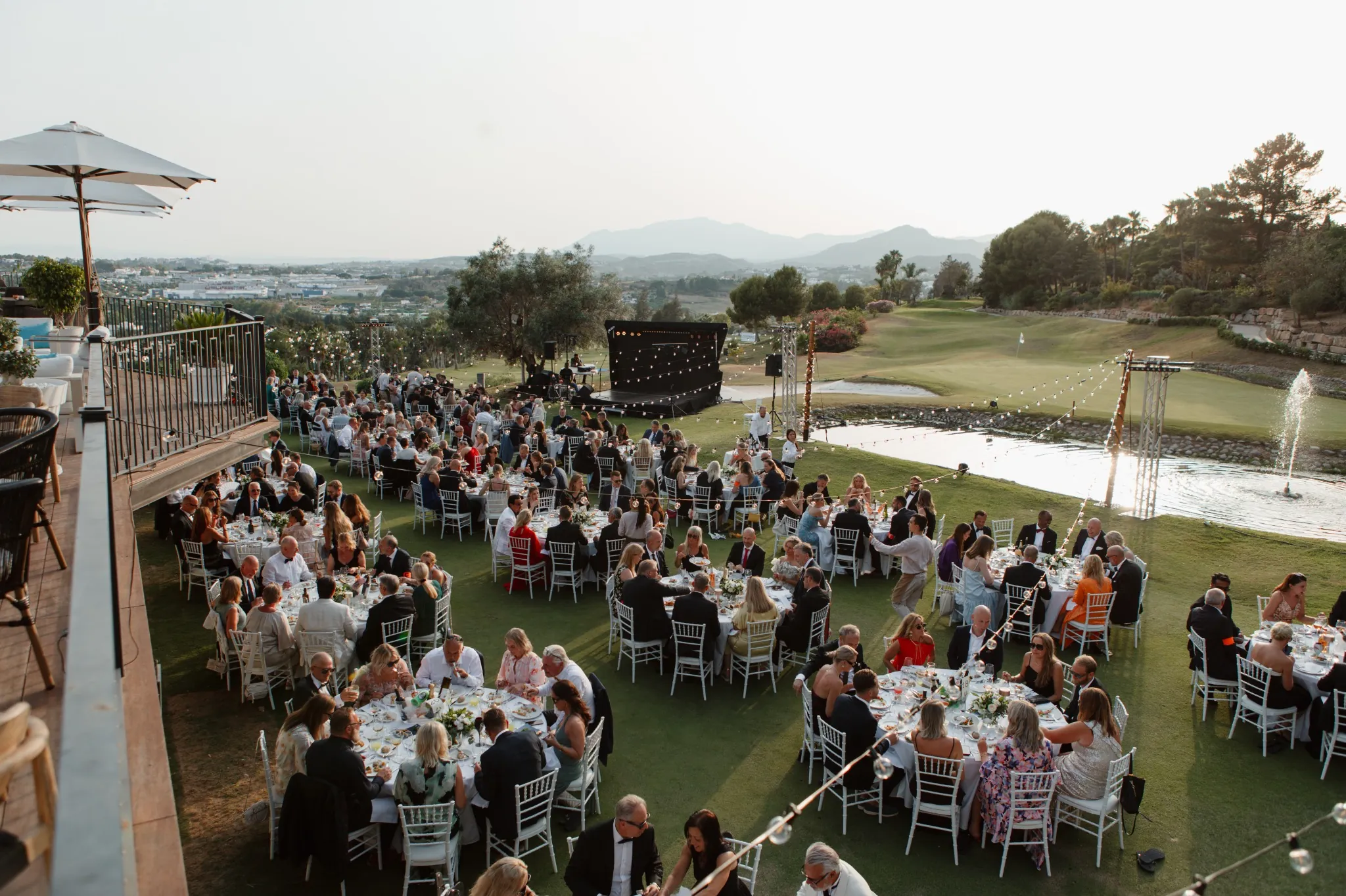 Round dinner tables on a lawn beside a pond and a stage at an outdoor evening event