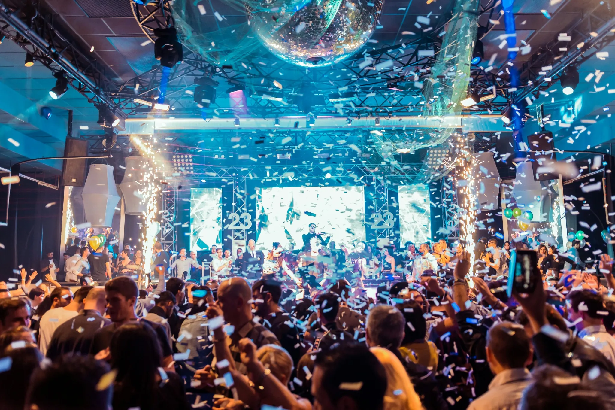 A crowd celebrating under confetti and a mirror ball in front of a stage at a large indoor event