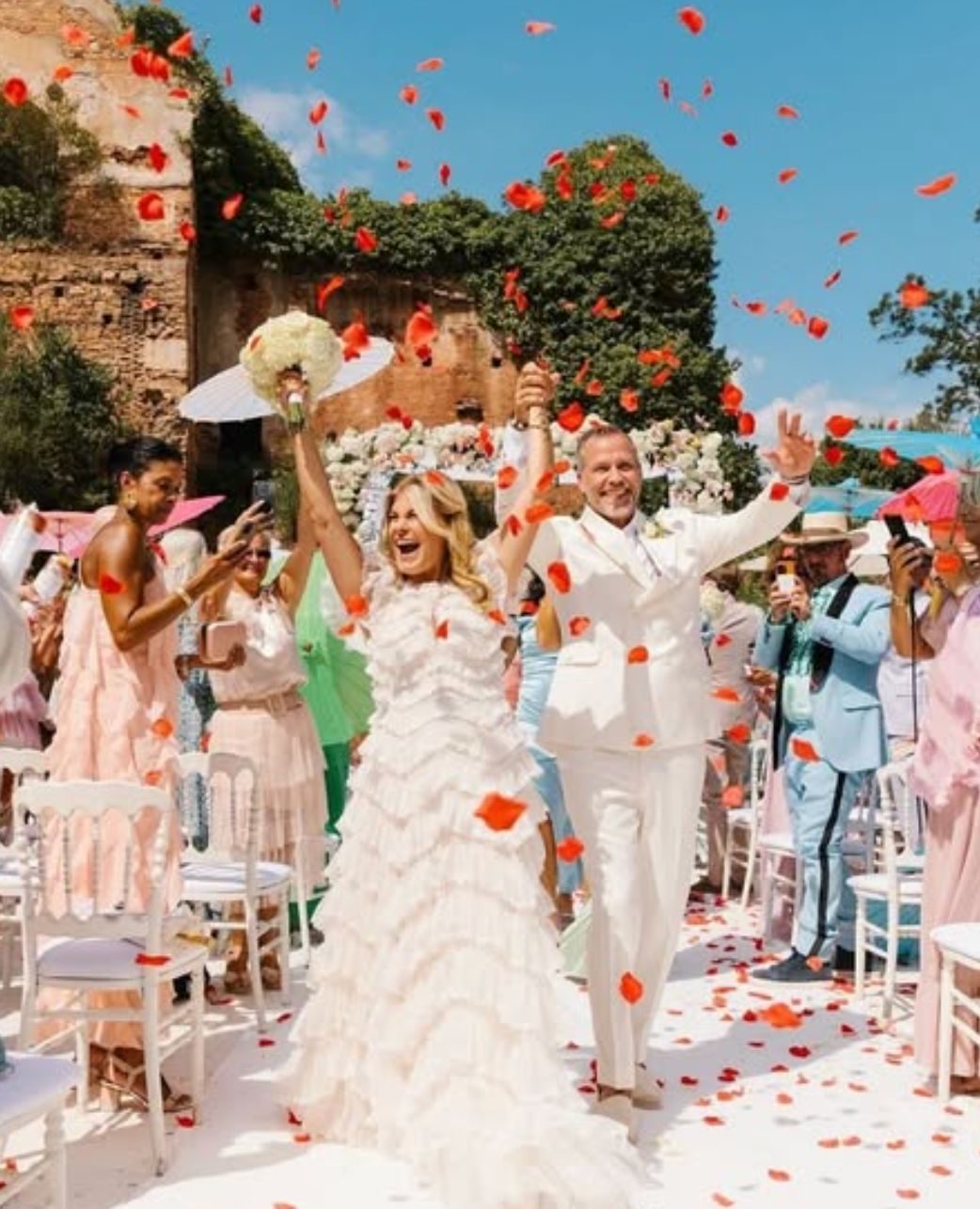 A couple walking back up the aisle under a shower of red petals at an outdoor wedding ceremony