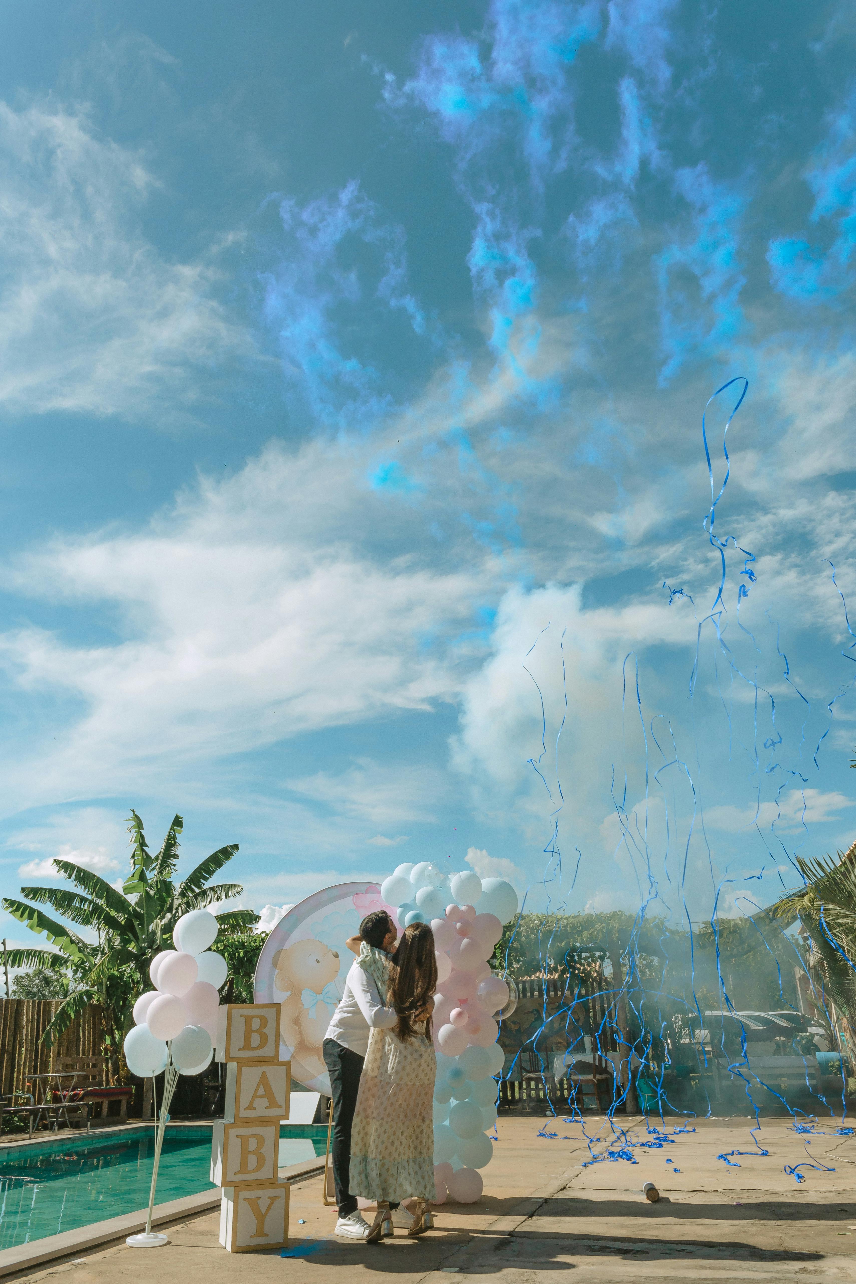 A couple beside a pool with balloons and 'BABY' blocks as blue smoke rises at a gender reveal