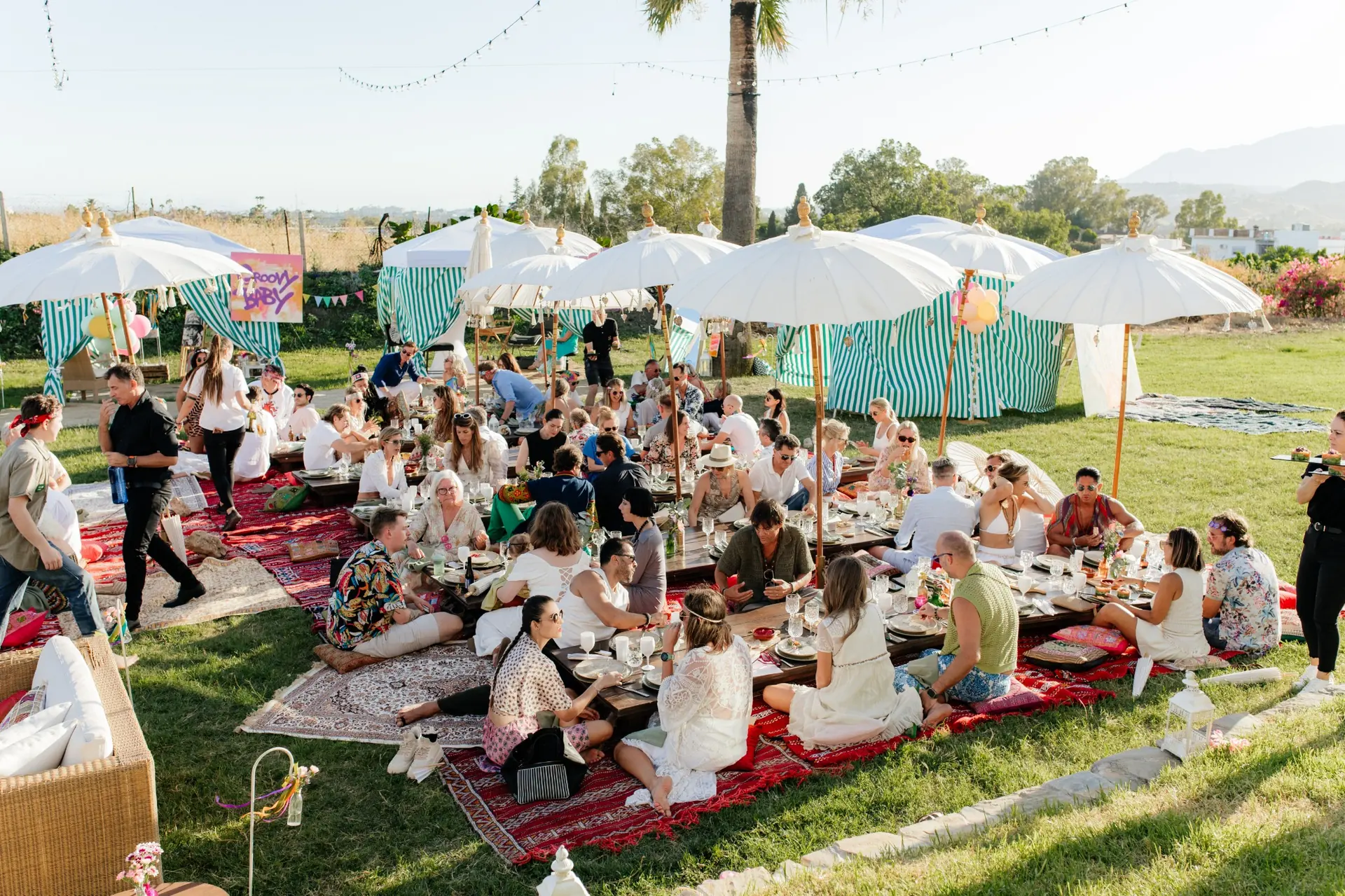 Guests at low tables under white parasols on a lawn at a daytime outdoor event